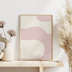 May include: A framed abstract print with pink and cream wavy shapes against a white background, displayed on a light wood shelf with two vases of dried flowers and a tall dried pampas grass plant.