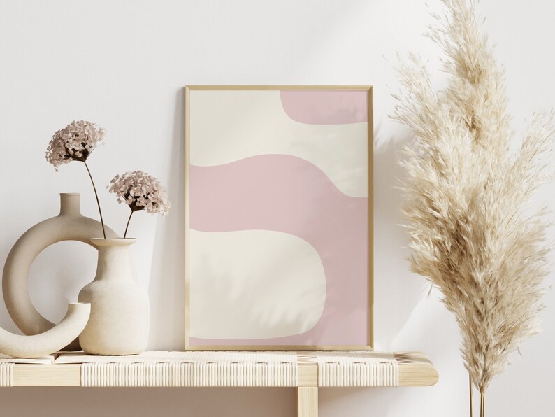 May include: A framed abstract print with pink and cream wavy shapes against a white background, displayed on a light wood shelf with two vases of dried flowers and a tall dried pampas grass plant.