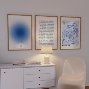 Op de afbeelding: Drie ingelijste prints op een witte muur. De linker print heeft een abstract ontwerp in blauw en wit. De middelste print heeft de tekst "Do all things with kindness" 18 keer herhaald. De rechter print heeft een abstract ontwerp in blauw en wit met het woord "GRATITUDE" onderaan.