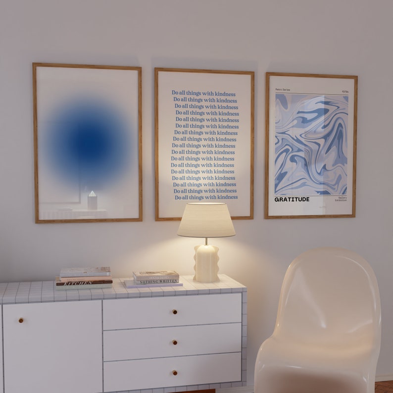 Op de afbeelding: Drie ingelijste prints op een witte muur. De linker print heeft een abstract ontwerp in blauw en wit. De middelste print heeft de tekst "Do all things with kindness" 18 keer herhaald. De rechter print heeft een abstract ontwerp in blauw en wit met het woord "GRATITUDE" onderaan.