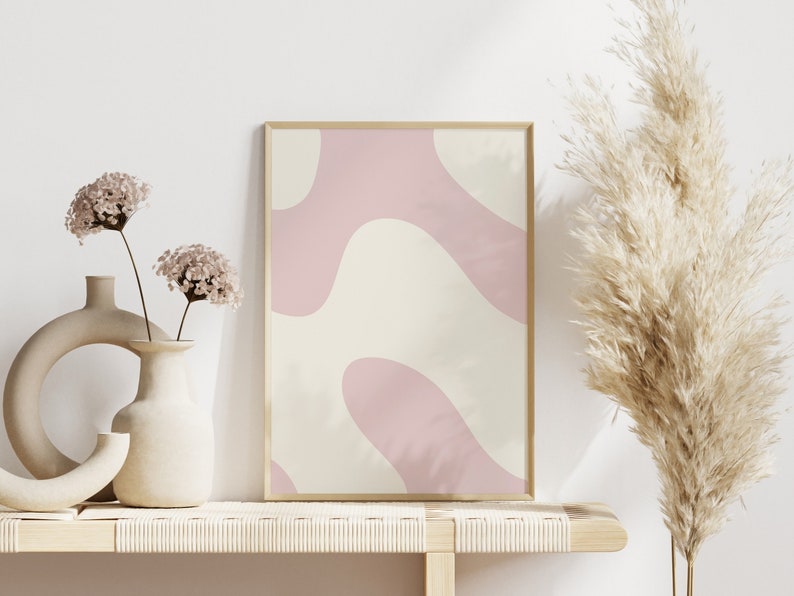 May include: A framed abstract print with pink and cream colored organic shapes. The print is displayed on a light wood shelf with two ceramic vases and dried flowers.