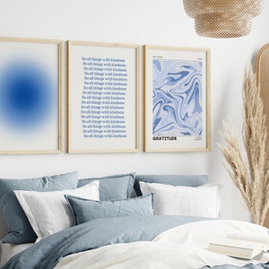 Op de afbeelding: Drie ingelijste prints op een witte muur boven een bed met blauwe beddengoed. De print aan de linkerkant is een blauwe gradi&euml;nt. De middelste print heeft de tekst "Do all things with kindness" 17 keer herhaald. De print aan de rechterkant heeft een blauw abstract ontwerp en de tekst "GRATITUDE".
