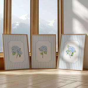 Pu&ograve; includere: Tre stampe artistiche incorniciate con fiori di ortensia ad acquerello. Ogni stampa ha uno sfondo a righe azzurre e bianche con un bordo azzurro. I fiori di ortensia sono azzurri con foglie verdi, su sfondo bianco.