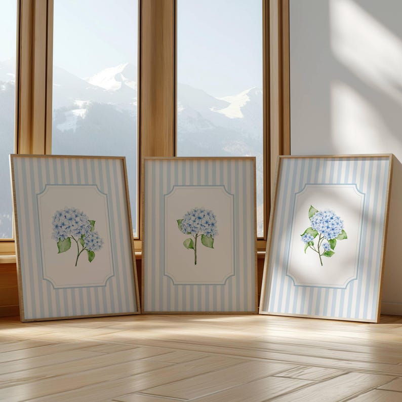Pu&ograve; includere: Tre stampe artistiche incorniciate con fiori di ortensia ad acquerello. Ogni stampa ha uno sfondo a righe azzurre e bianche con un bordo azzurro. I fiori di ortensia sono azzurri con foglie verdi, su sfondo bianco.