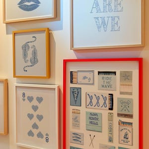 May include: A collection of framed art pieces with a variety of designs. The art includes blue lips, cheetahs, playing cards, and matchbox art with phrases like "Ride the Wave" and "Aloha, Howdy, Hello." The frames are in various colors, including red and gold.