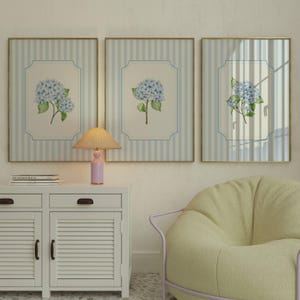 Pu&ograve; includere: Tre opere d'arte incorniciate con sfondi a righe blu e bianche e cornici dorate. Ognuna presenta un dipinto ad acquerello di ortensie blu con foglie verdi. Una piccola lampada e un mobile bianco sono in primo piano.