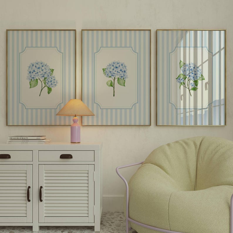 Pu&ograve; includere: Tre opere d'arte incorniciate con sfondi a righe blu e bianche e cornici dorate. Ognuna presenta un dipinto ad acquerello di ortensie blu con foglie verdi. Una piccola lampada e un mobile bianco sono in primo piano.