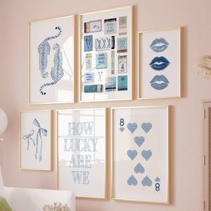 May include: A collection of framed art prints with gold frames. The prints feature blue and white designs, including leopards, lips, playing cards, and the text "How Lucky Are We". The art is displayed on a pink wall.