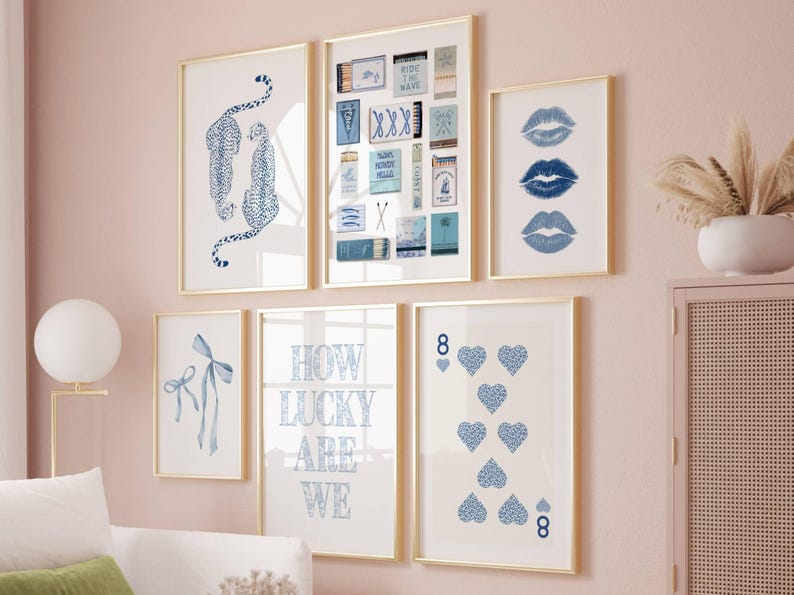 May include: A collection of framed art prints with gold frames. The prints feature blue and white designs, including leopards, lips, playing cards, and the text "How Lucky Are We". The art is displayed on a pink wall.