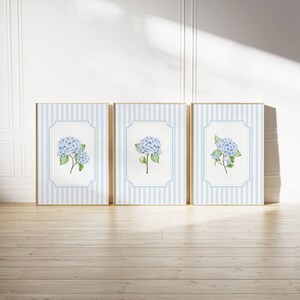 Pu&ograve; includere: Tre stampe artistiche incorniciate con dipinti ad acquerello di fiori di ortensia blu. Ogni stampa ha uno sfondo a righe blu chiaro e bianco con un bordo bianco. Le cornici sono in legno chiaro, su un pavimento in legno.