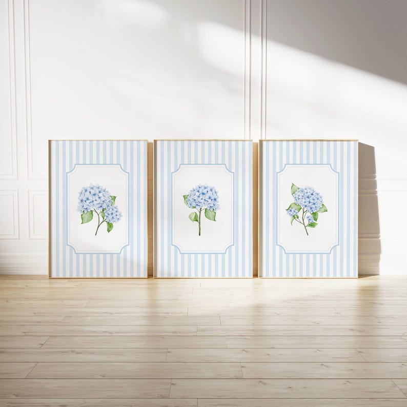 Pu&ograve; includere: Tre stampe artistiche incorniciate con dipinti ad acquerello di fiori di ortensia blu. Ogni stampa ha uno sfondo a righe blu chiaro e bianco con un bordo bianco. Le cornici sono in legno chiaro, su un pavimento in legno.
