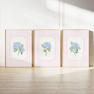 May include: Three framed art prints featuring watercolor illustrations of blue hydrangea flowers. The prints have a pink and white striped background with a decorative pink border. The frames are a light wood color.