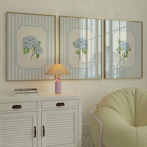 Pu&ograve; includere: Tre quadri incorniciati con bordi dorati, ognuno con un'illustrazione di ortensia blu su sfondo a righe. Una piccola lampada rosa &egrave; appoggiata su un mobile bianco sotto le opere d'arte. Sono visibili anche una pila di libri e una poltrona sacco gialla.