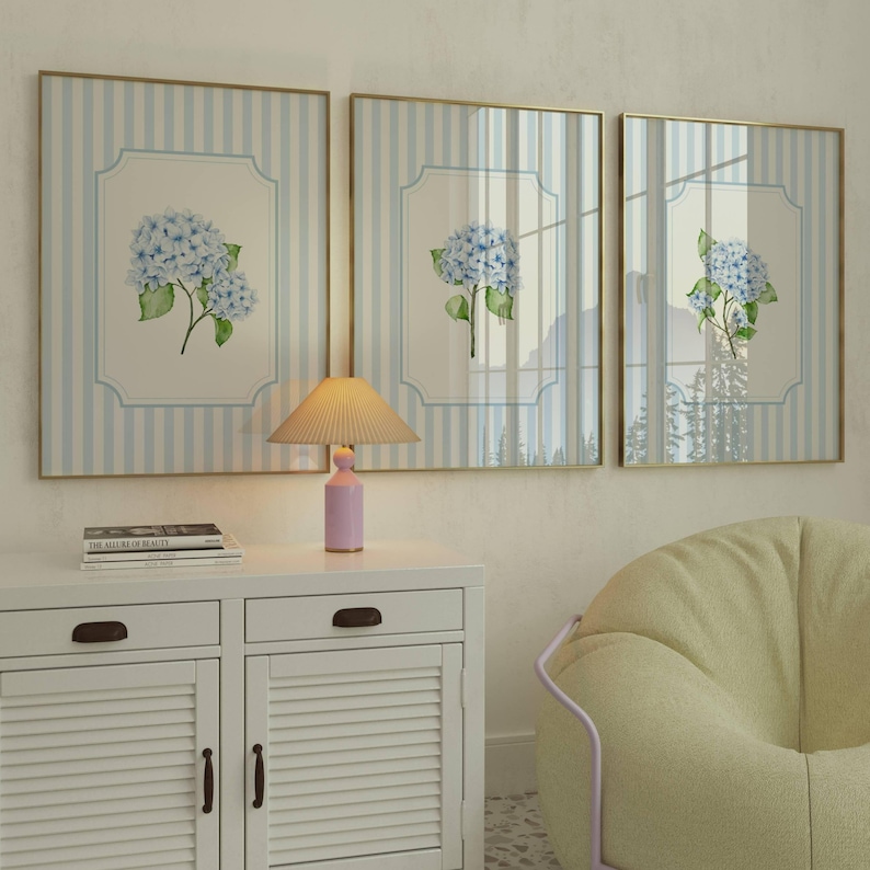 Pu&ograve; includere: Tre quadri incorniciati con bordi dorati, ognuno con un'illustrazione di ortensia blu su sfondo a righe. Una piccola lampada rosa &egrave; appoggiata su un mobile bianco sotto le opere d'arte. Sono visibili anche una pila di libri e una poltrona sacco gialla.