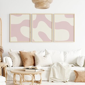 May include: Three framed abstract art prints with pink and cream colored organic shapes. The prints are hanging on a white wall above a white couch with brown throw pillows and a woven coffee table.