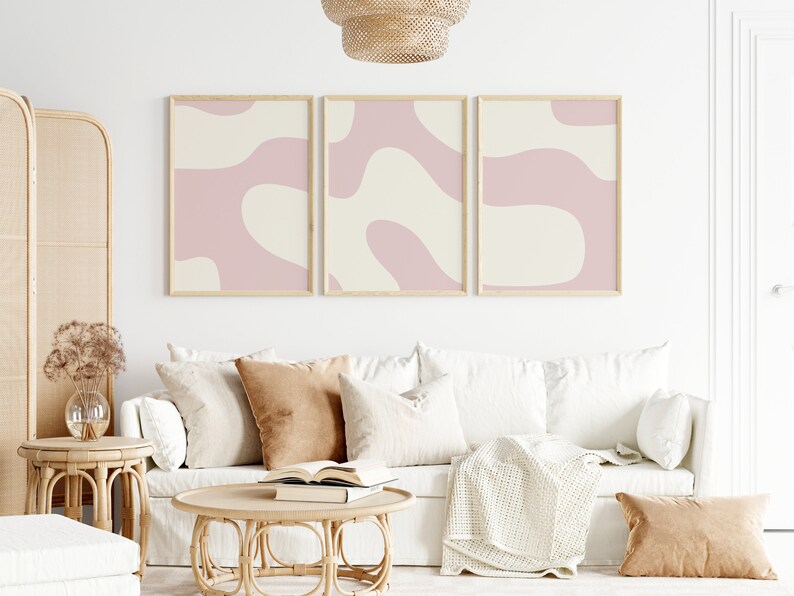 May include: Three framed abstract art prints with pink and cream colored organic shapes. The prints are hanging on a white wall above a white couch with brown throw pillows and a woven coffee table.