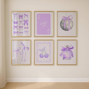May include: Six framed art prints with gold frames. The prints feature watercolor illustrations of bows, a playing card with text, a disco ball with a bow, cherries, and a perfume bottle, all in shades of purple. The prints are displayed on a cream-colored wall.