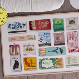 May include: A collection of 16 matchbox labels featuring various designs, including a Jeep, a cat, a bowling ball, a strawberry, a cityscape, and a palm tree. The labels are arranged in a grid pattern on a white background.