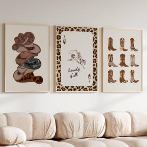 Pode incluir: Três impressões emolduradas com tema western. A impressão da esquerda apresenta uma pilha de chapéus de cowboy marrons com um design de estrela. A impressão central tem um fundo de estampa de leopardo com um ás de paus e o texto "Howdy y'all". A impressão da direita apresenta uma coleção de botas de cowboy marrons.