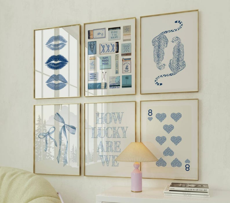 May include: Six framed art prints with gold frames. The prints feature blue lips, matchboxes, cheetahs, a bow, the text "HOW LUCKY ARE WE", and playing card hearts. The art is displayed on a white wall.