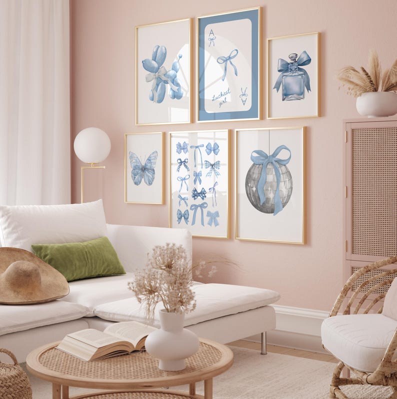 May include: A living room interior with a white sectional sofa, a wicker chair, and a coffee table. The wall is painted a light pink color and features a gallery wall of framed prints with blue and white watercolor illustrations of bows, butterflies, and a disco ball. The text "Luckiest girl" is visible on one of the prints.