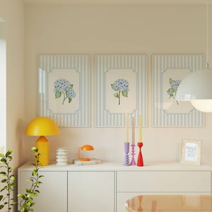 Pu&ograve; includere: Tre stampe floreali incorniciate con fiori di ortensia blu sono appese sopra un mobile bianco. Una lampada a fungo gialla e una lampada da scrivania arancione sono sul mobile, insieme a candele decorative e una piccola stampa incorniciata con la scritta "I miss you bye".