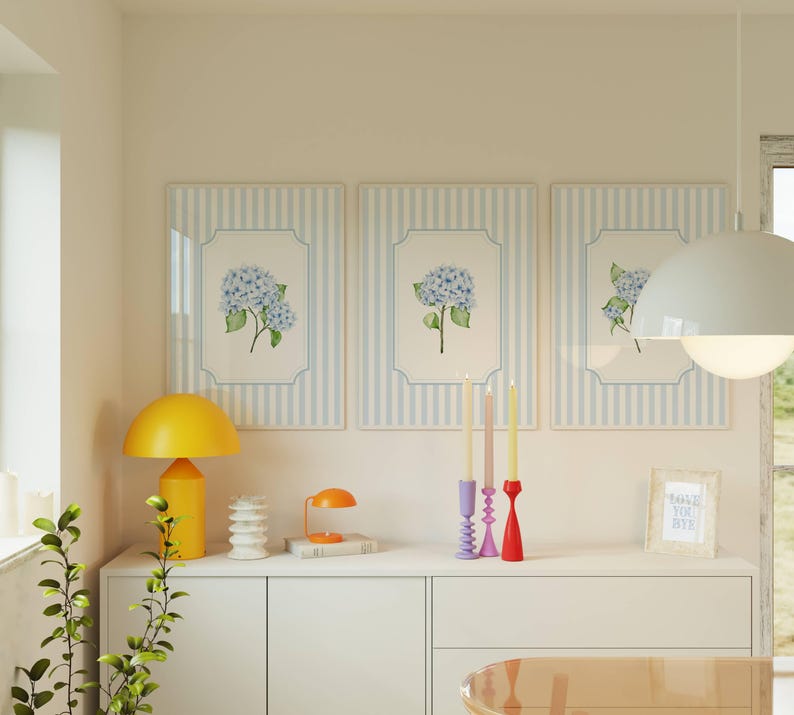 Pu&ograve; includere: Tre stampe floreali incorniciate con fiori di ortensia blu sono appese sopra un mobile bianco. Una lampada a fungo gialla e una lampada da scrivania arancione sono sul mobile, insieme a candele decorative e una piccola stampa incorniciata con la scritta "I miss you bye".