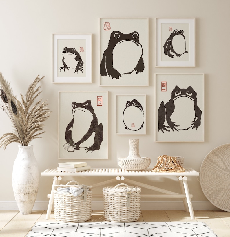 May include: A white wooden bench with two white wicker baskets underneath. There are five framed black and white prints of frogs on the wall above the bench. The prints are arranged in a symmetrical pattern. A vase with dried pampas grass sits on the floor to the left of the bench.