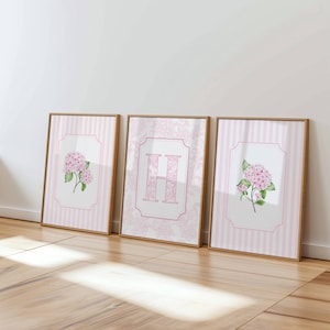 May include: Three framed wall art prints with light wood frames. The outer two prints feature pink hydrangea illustrations on a white background with pink vertical stripes. The center print displays a large pink letter H with a floral pattern.