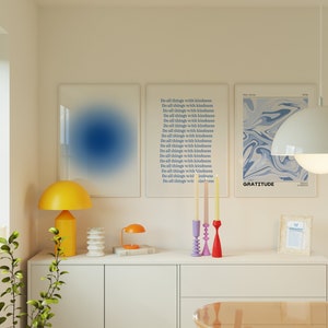 Op de afbeelding: Drie ingelijste prints met blauwe en witte abstracte ontwerpen. De prints hangen aan een witte muur boven een witte commode met een gele lamp, een kleine tafellamp en een kandelaar met drie kaarsen. De tekst op de prints luidt "Do all things with kindness" en "Gratitude".