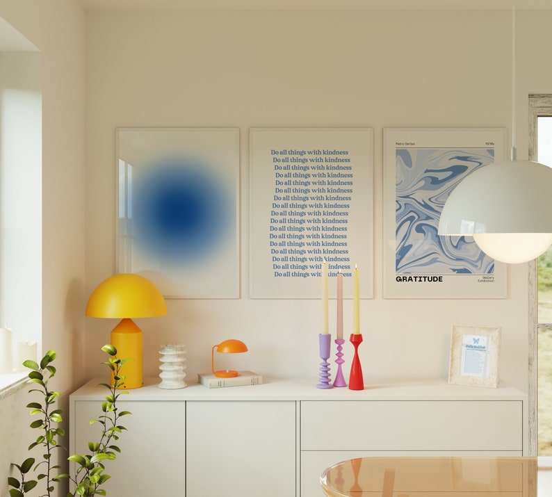 Op de afbeelding: Drie ingelijste prints met blauwe en witte abstracte ontwerpen. De prints hangen aan een witte muur boven een witte commode met een gele lamp, een kleine tafellamp en een kandelaar met drie kaarsen. De tekst op de prints luidt "Do all things with kindness" en "Gratitude".