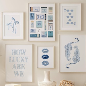 May include: A collection of framed art prints in shades of blue and white. The prints feature various designs, including bows, playing cards, matchbox art, lips, and cheetahs. The phrase "HOW LUCKY ARE WE" is also displayed.
