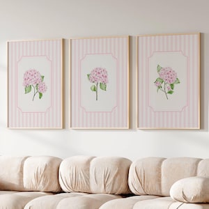 May include: Three framed floral prints showcasing pink hydrangea flowers. Each print has a pink and white striped background with a white, scalloped border around the floral image. The frames are gold-coloured.