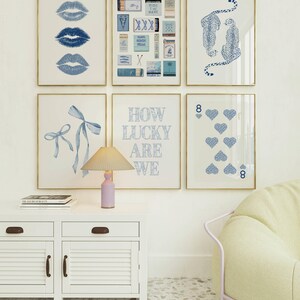 May include: A gallery wall featuring six framed art prints. The prints showcase various blue designs, including lips, matchboxes, cheetahs, bows, playing cards, and the text "HOW LUCKY ARE WE". A white cabinet with a lamp and books sits below.
