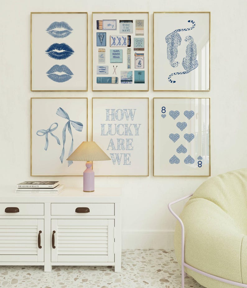 May include: A gallery wall featuring six framed art prints. The prints showcase various blue designs, including lips, matchboxes, cheetahs, bows, playing cards, and the text "HOW LUCKY ARE WE". A white cabinet with a lamp and books sits below.