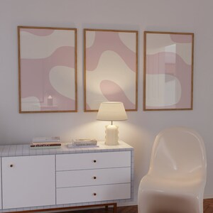 May include: Three framed abstract art prints with pink and white organic shapes, a white lamp with a white lampshade, a white dresser with three drawers, and a white chair.
