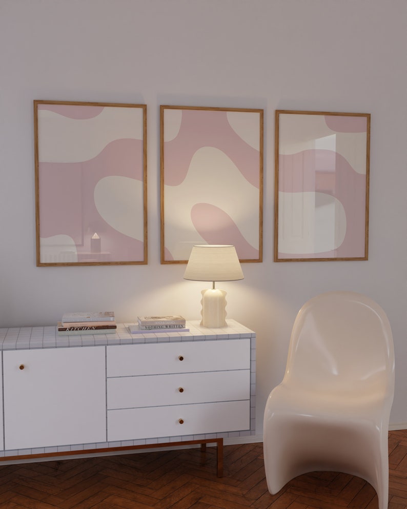May include: Three framed abstract art prints with pink and white organic shapes, a white lamp with a white lampshade, a white dresser with three drawers, and a white chair.