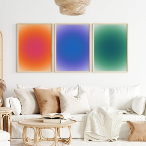Peut inclure: Trois impressions abstraites d&eacute;grad&eacute;es encadr&eacute;es dans un salon. Les impressions sont d'un orange, bleu et vert vifs. Les impressions sont accroch&eacute;es &agrave; un mur blanc au-dessus d'un canap&eacute; blanc avec des coussins et une couverture.
