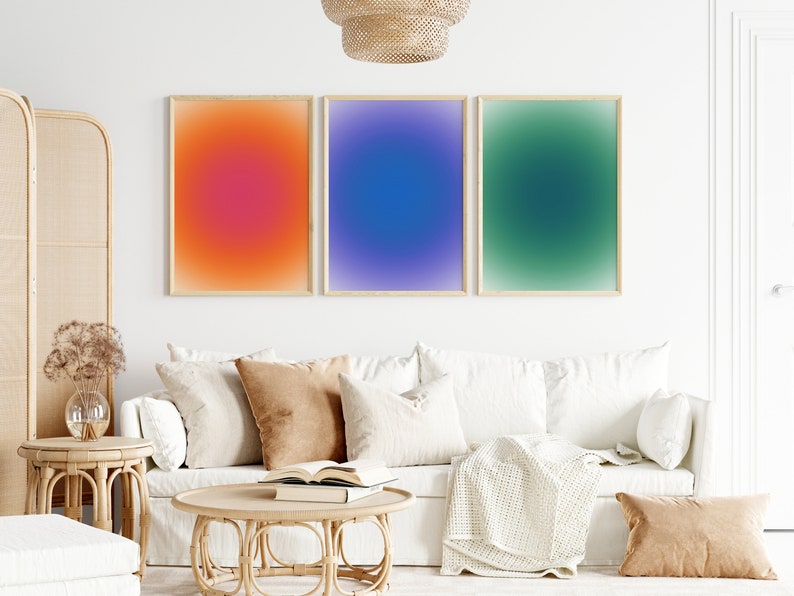 Peut inclure: Trois impressions abstraites d&eacute;grad&eacute;es encadr&eacute;es dans un salon. Les impressions sont d'un orange, bleu et vert vifs. Les impressions sont accroch&eacute;es &agrave; un mur blanc au-dessus d'un canap&eacute; blanc avec des coussins et une couverture.