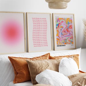 K&ouml;nnte beinhalten: Drei gerahmte Drucke an einer wei&szlig;en Wand &uuml;ber einem Bett mit Kissen. Der linke Druck hat einen rosa Farbverlauf. Der mittlere Druck hat den Text "Good things are coming" 17 Mal in rosa wiederholt. Der rechte Druck hat ein farbenfrohes abstraktes Design und den Text "ABUNDANCE" unten.