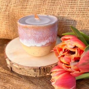 Op de afbeelding: Een witte keramische kaars pot met een houten lont, die op een houten schijf staat, naast een boeket oranje en gele tulpen.