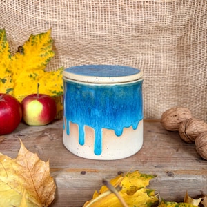 以下が含まれることがあります： 蓋付きのセラミックジャー。鮮やかな青色の釉薬が、まだらの白いベースに滴り落ちています。円筒形のジャーは木製の表面に置かれ、秋の葉やリンゴに囲まれています。