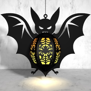 May include: A black bat silhouette with a glowing orange and yellow spiderweb sphere hanging from its center. The bat is hanging from a string against a white concrete wall.