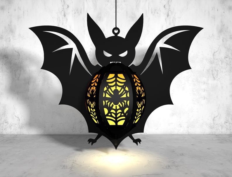 May include: A black bat silhouette with a glowing orange and yellow spiderweb sphere hanging from its center. The bat is hanging from a string against a white concrete wall.