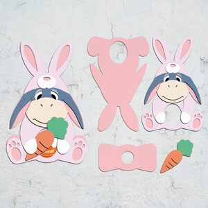 May include: A pink bunny costume with a gray donkey face, holding a carrot. The costume is made of felt and has a hole for a child's head. There is also a separate pink bunny head and a pink bow tie.