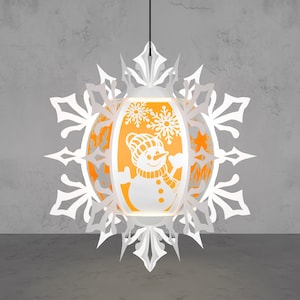 May include: A white paper lantern with a snowman design, surrounded by a snowflake-shaped frame. The lantern has an orange interior, and is hanging from a black cord. The snowman is wearing a hat and scarf.