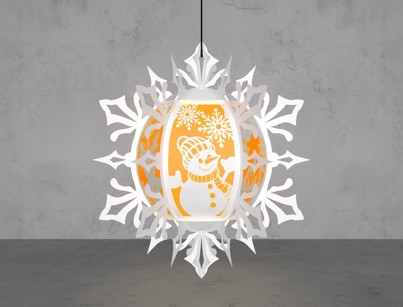 May include: A white paper lantern with a snowman design, surrounded by a snowflake-shaped frame. The lantern has an orange interior, and is hanging from a black cord. The snowman is wearing a hat and scarf.