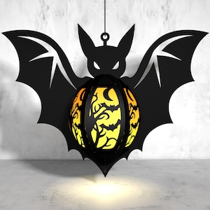 May include: A black bat-shaped lantern with a yellow and orange patterned sphere in the center. The sphere has a black silhouette of a tree and bats.