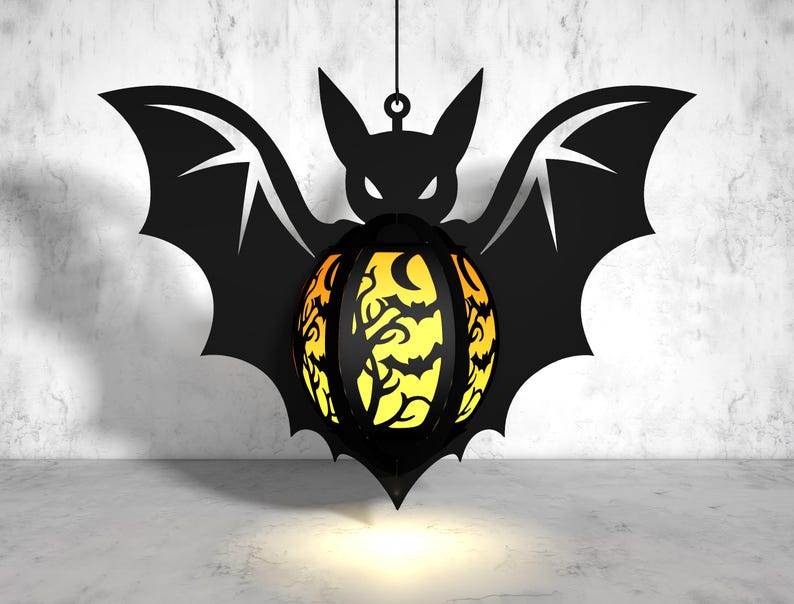 May include: A black bat-shaped lantern with a yellow and orange patterned sphere in the center. The sphere has a black silhouette of a tree and bats.