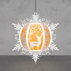 May include: A white paper lantern with a deer silhouette and tree design, surrounded by snowflake cutouts. The lantern has an orange interior and hangs from a black cord. The design is intricate and festive.